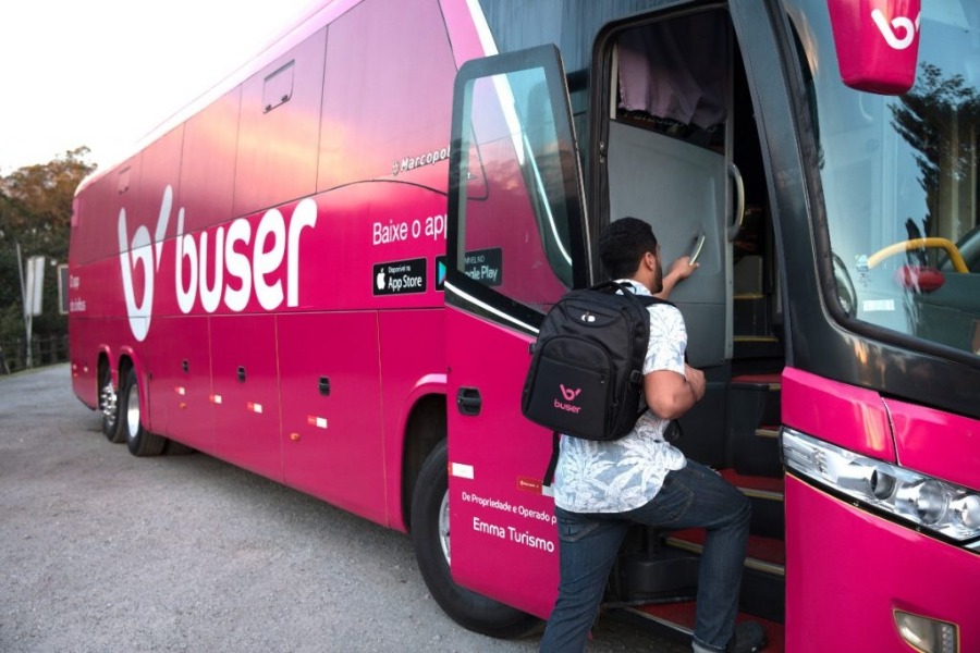 na imagem existe um ônibus rosa da Buser ao fundo, com um passageiro homem de mochila subindo suas escadas, pois ele prefere viajar de ônibus a viajar de carro ou avião.